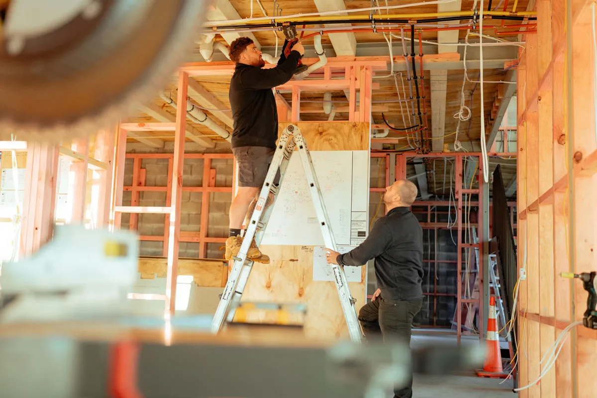 Two construction workers install piping in a wooden framework structure; one on ladder, one steadying it.