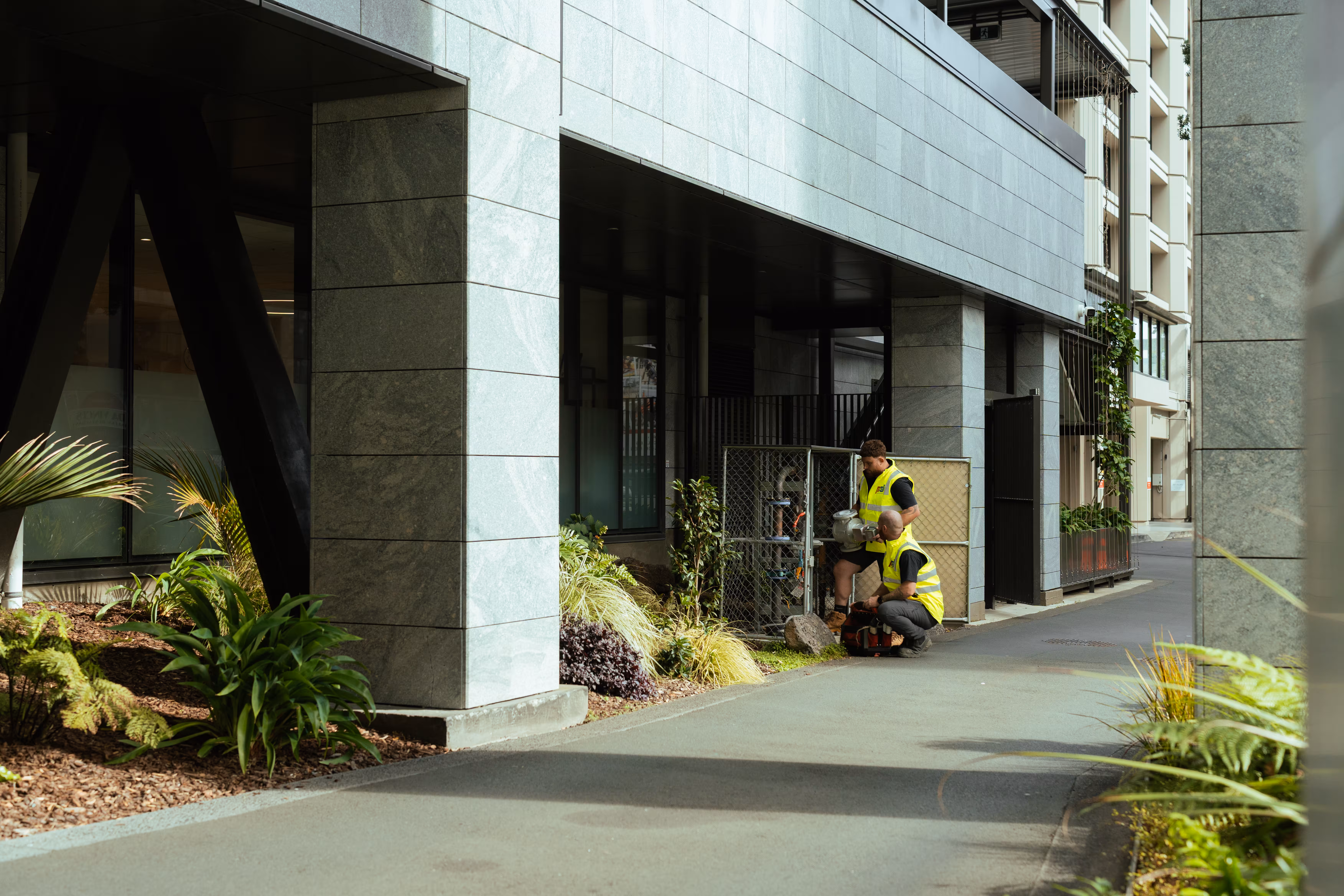 Two workers in yellow vests inspecting gas meters next to modern building with plants around.