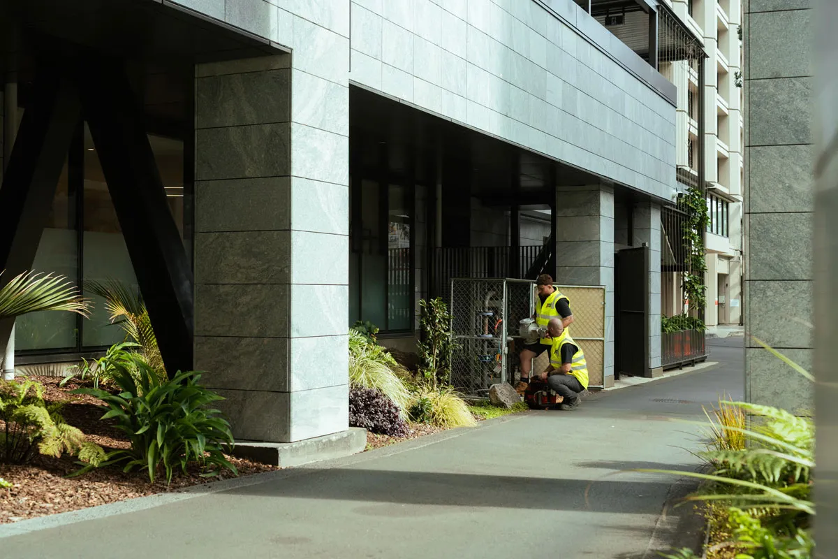 Two workers in yellow vests inspecting gas meters next to modern building with plants around.