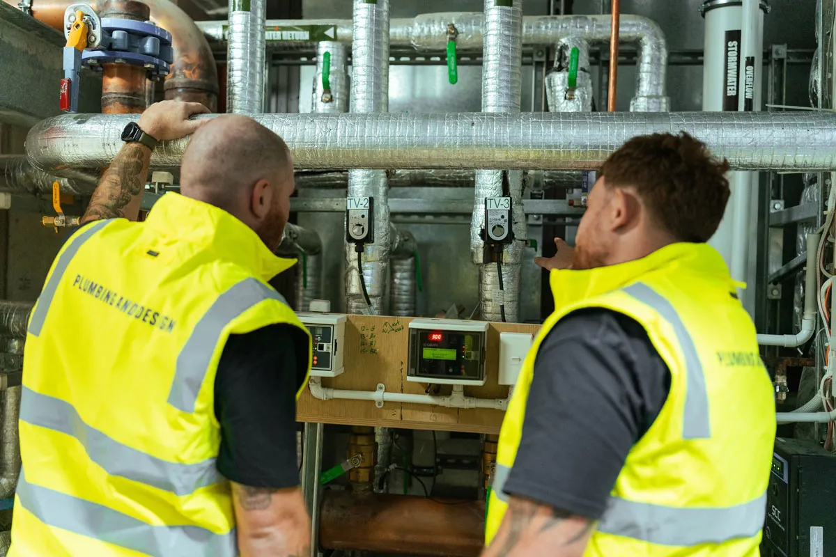 Two technicians in yellow vests inspecting and pointing at industrial plumbing pipes and control meters.