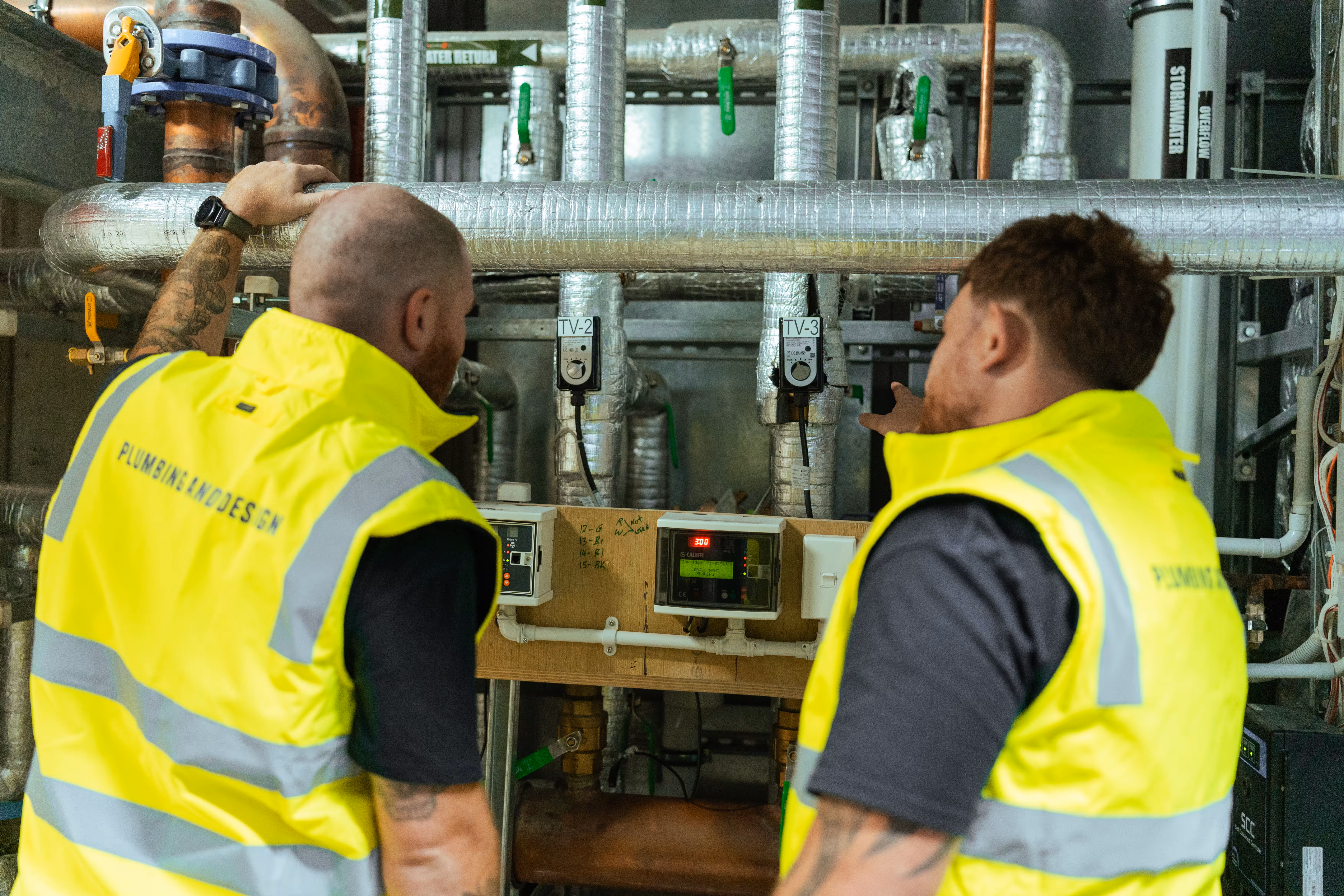 Two technicians in yellow vests inspecting and pointing at industrial plumbing pipes and control meters.