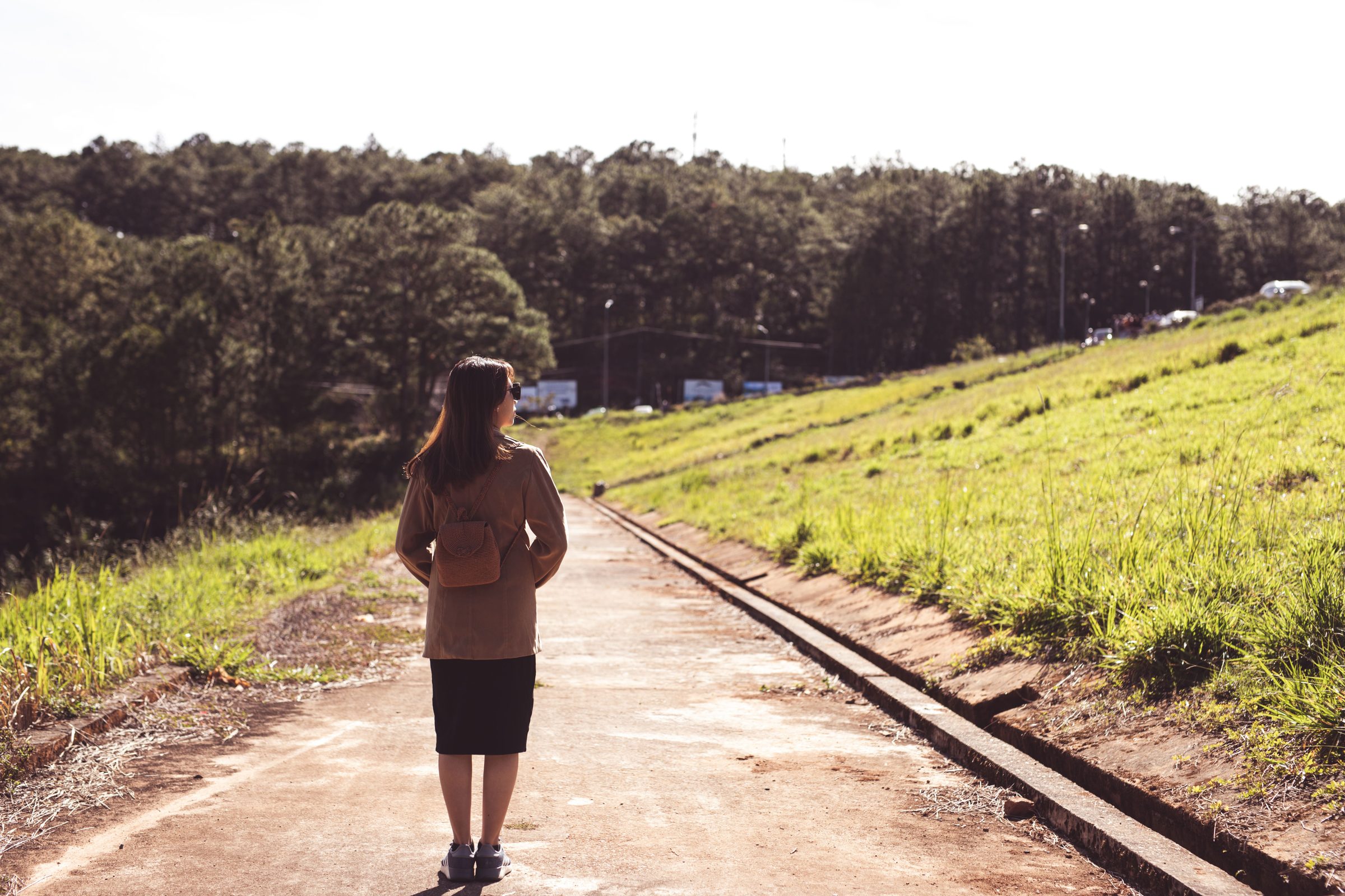 A woman walking outdoors along a sunlit, tree-lined path