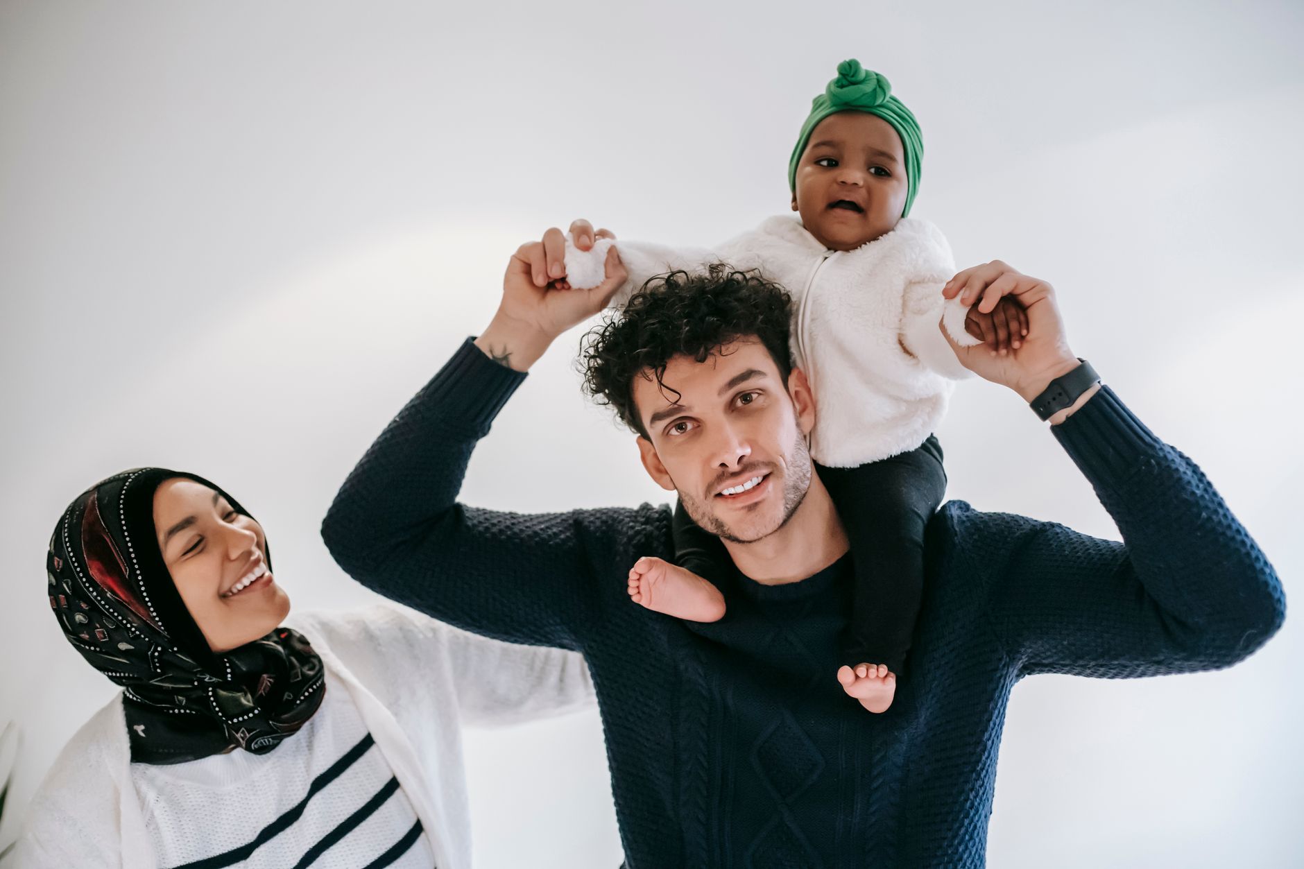 Positive dad enjoying fatherhood with girl on shoulders near Muslim mother in traditional hijab against white background