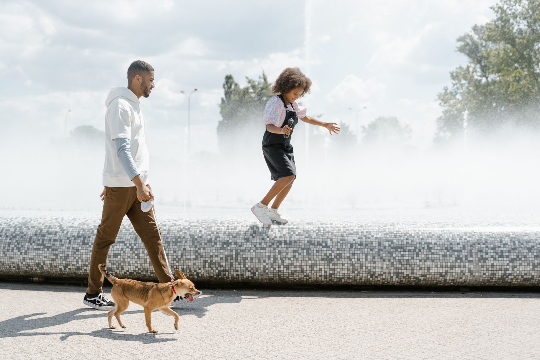 A father and daughter walking their dog by a lively water fountain in the park.