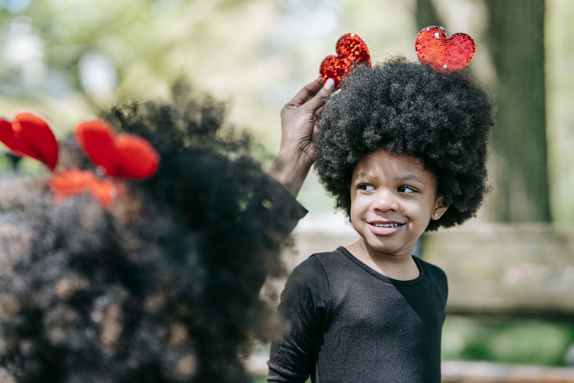A girl with afro hair smiles as her parent places heart-shaped accessories on her head outdoors.