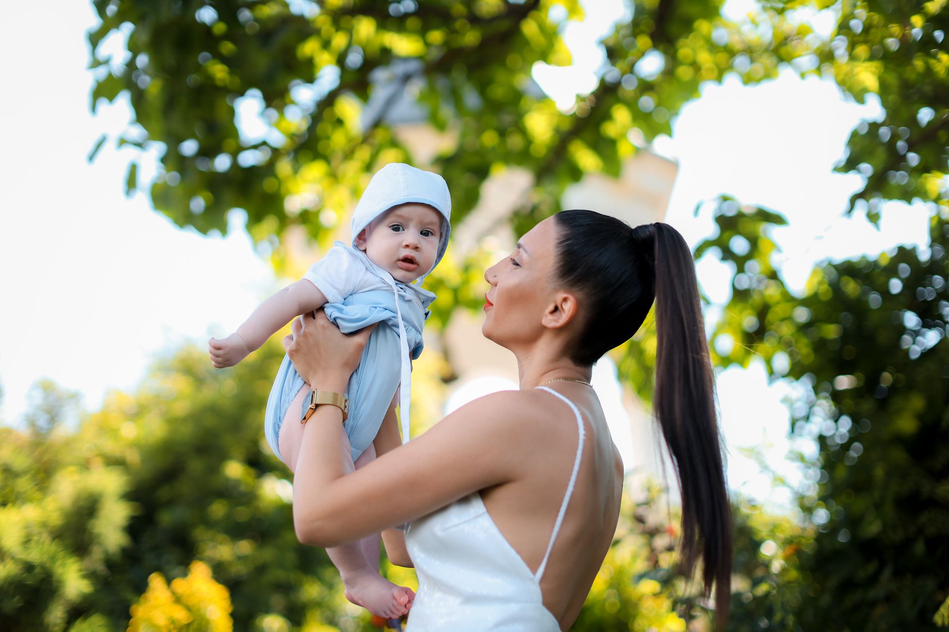 A joyful mother holding her baby under a green leafy tree on a sunny day.