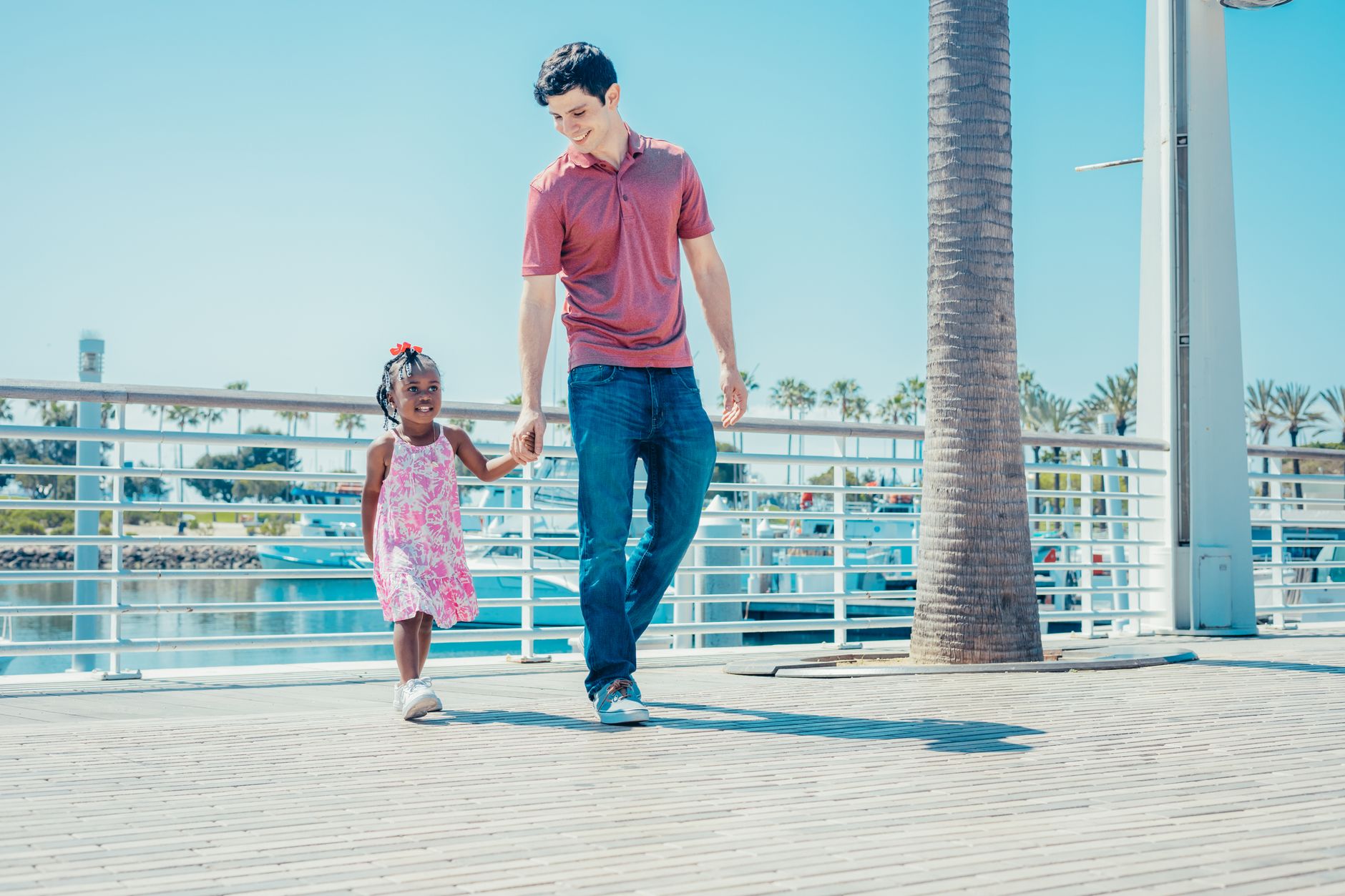 A dad and daughter enjoy a sunny walk by the marina, holding hands and smiling.