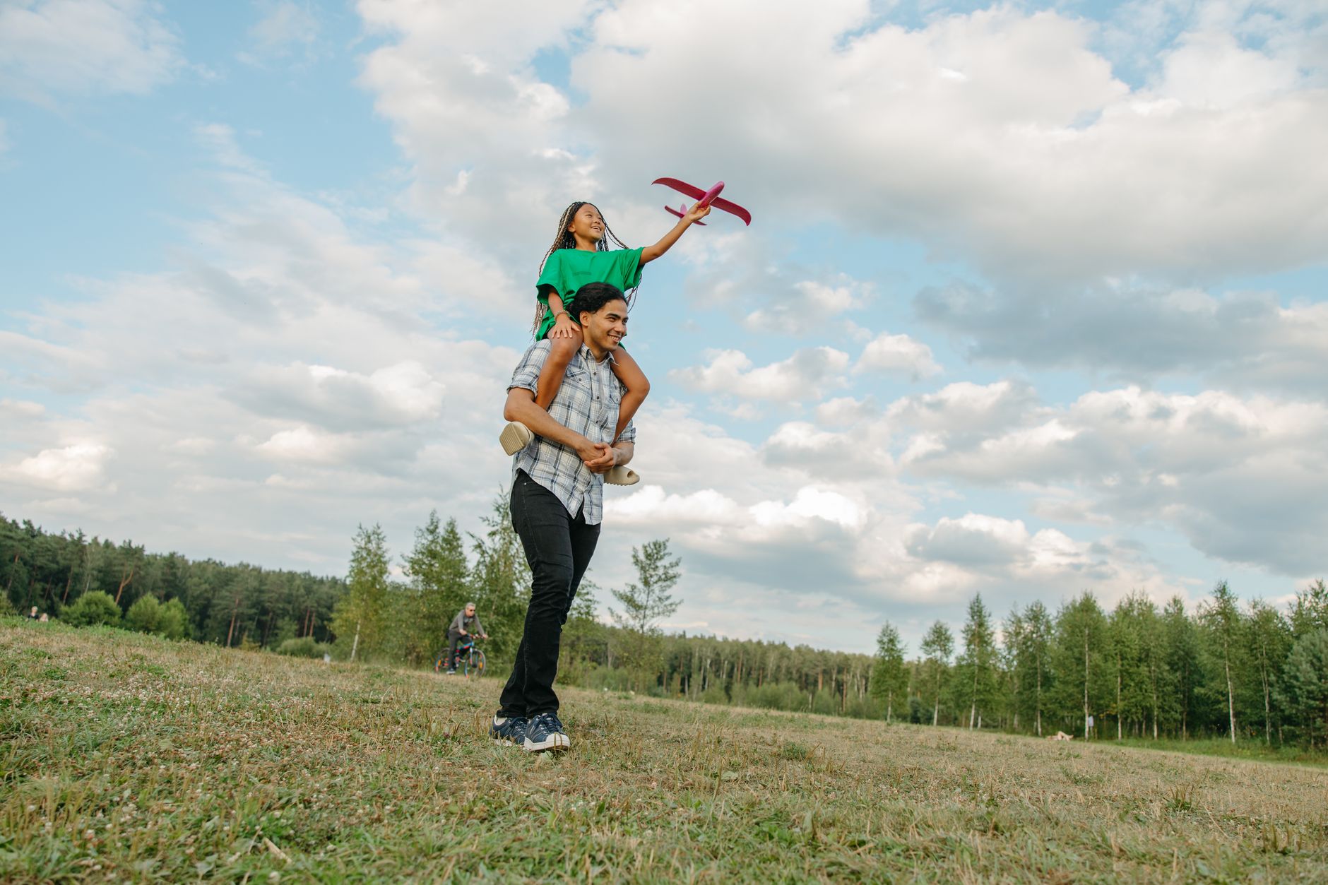 A joyful father carrying his daughter on shoulders in a sunny park playing with a toy plane.