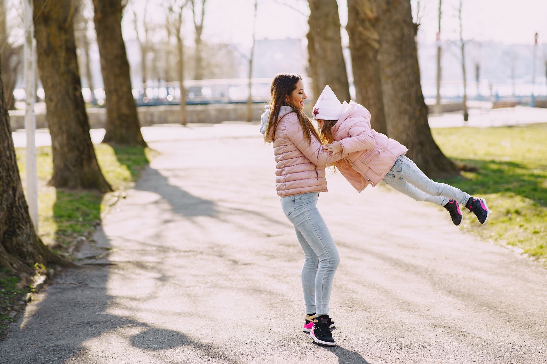 Side view of full body woman and female kid in warm clothes spining around on asphalt path near trees while walking in s