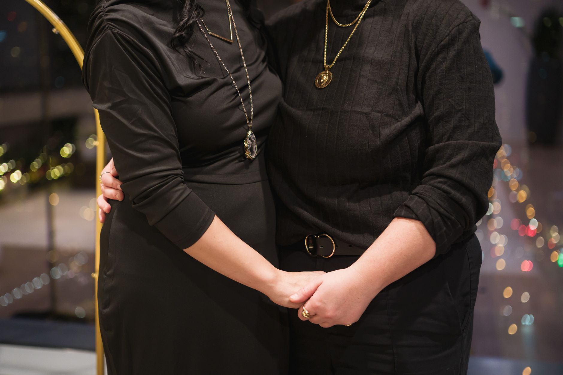 Two people hold hands in a warmly lit indoor setting wearing black outfits and necklaces.