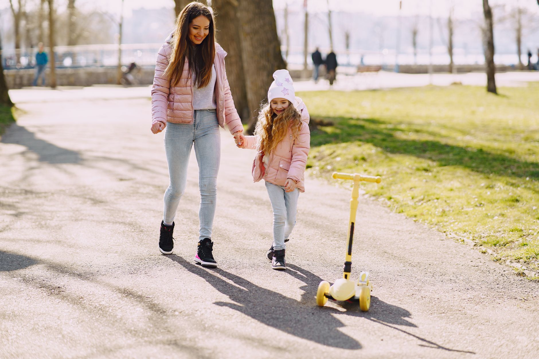 Full length female and little girl in warm clothes holding hands while walking and playing on walkway in city park near