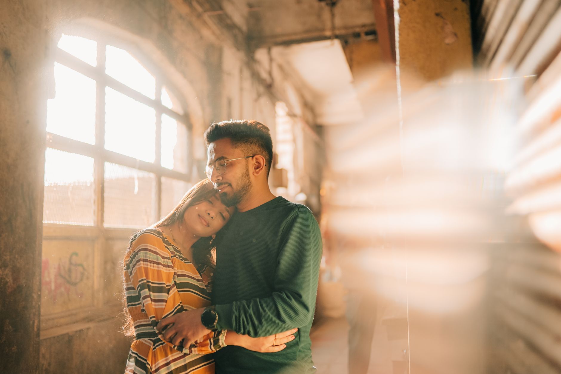 A loving couple hugs warmly under sunlight in a rustic indoor setting in Vadodara, India.
