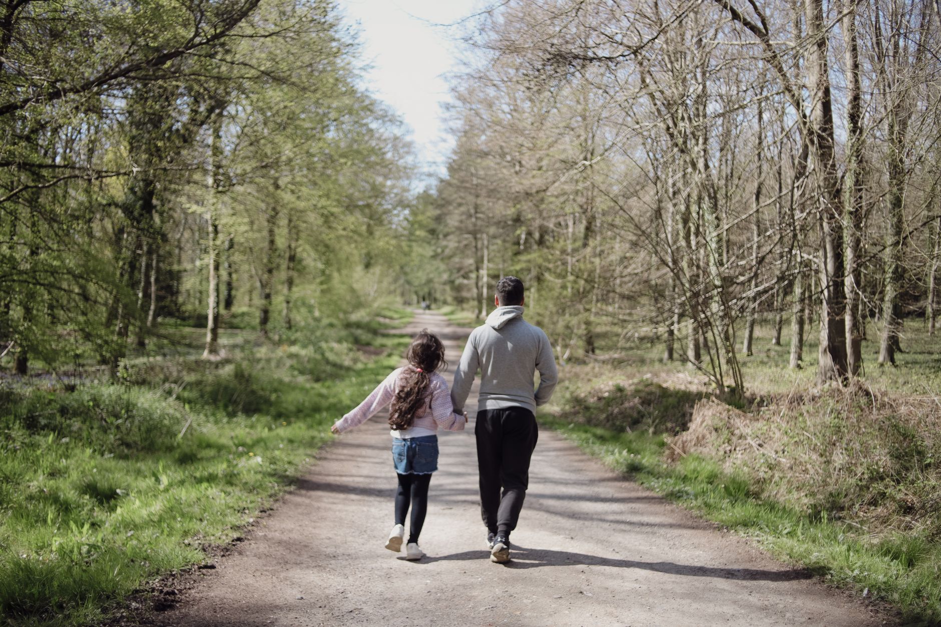 A father and daughter walk hand in hand on a scenic forest path, enjoying a sunny day.