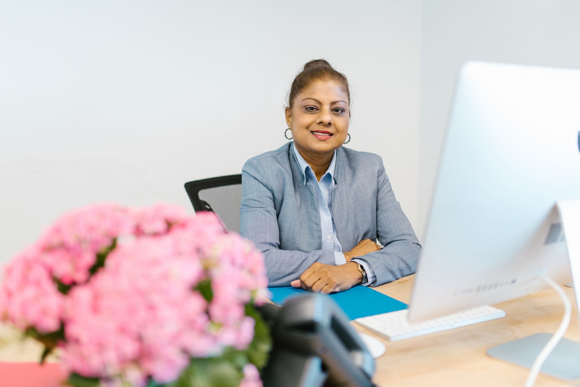 A smiling businesswoman sitting at her desk with flowers, wearing a blue suit in an office environment.