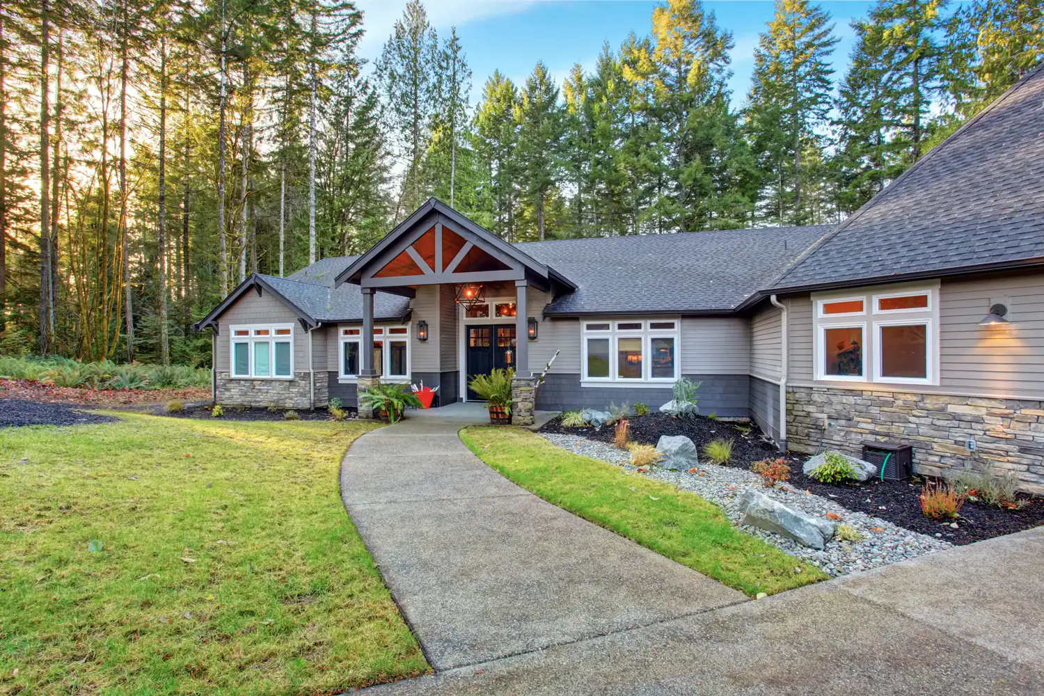 Modern house with stone and wood siding, a front porch, pathway, and forested background.