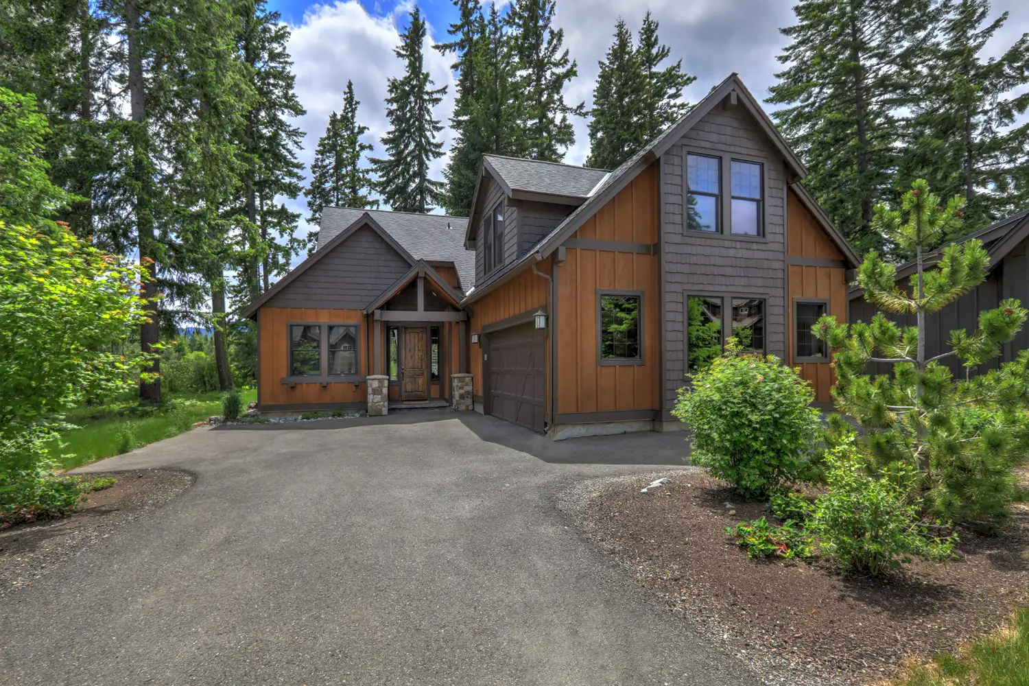 Modern wood and dark gray house with a driveway and surrounding trees and bushes.