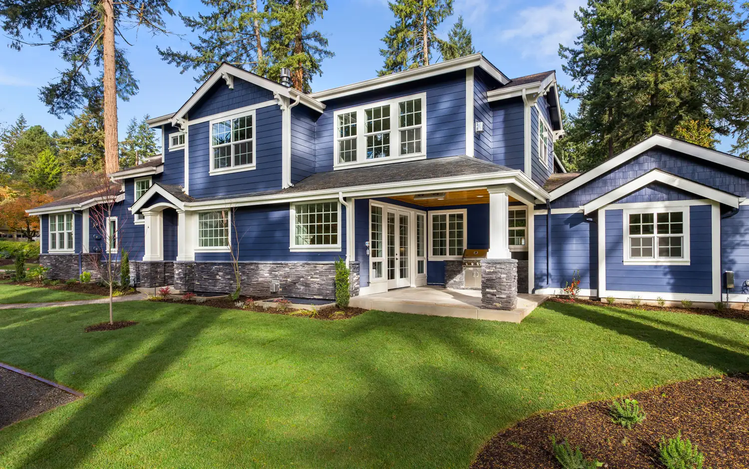 Two-story blue house with white trim, stone foundation, patio, and green lawn surrounded by trees.