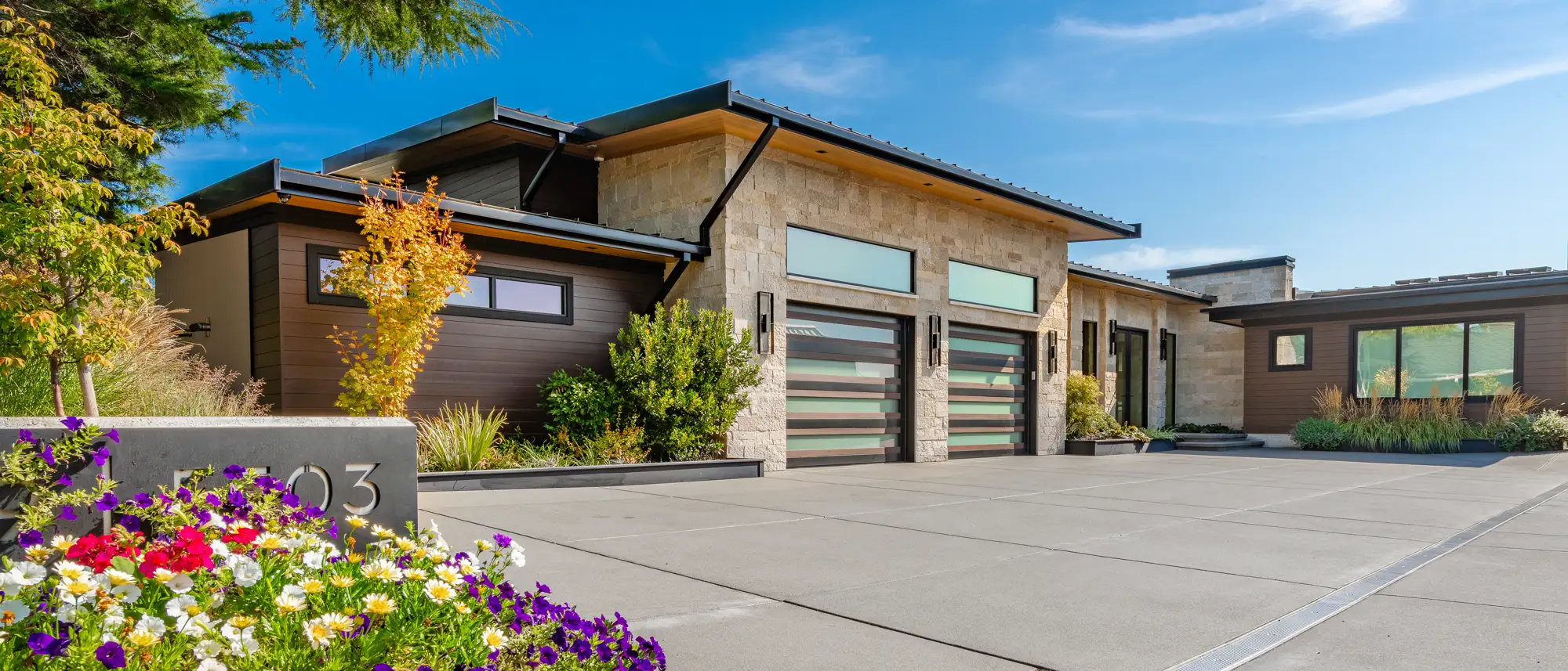 Modern house with dual garages, stone and wood exterior, surrounded by greenery and colorful flowers.