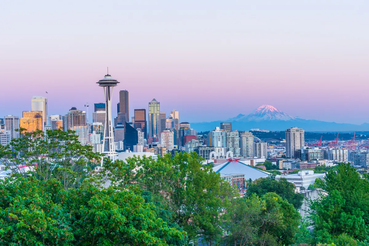 Seattle skyline with the Space Needle, green trees, and Mount Rainier under a pink sky at sunset.