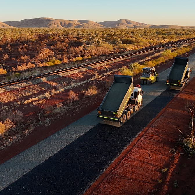 Two dump trucks and a road roller paving asphalt on a remote highway near dry hills and railway tracks.