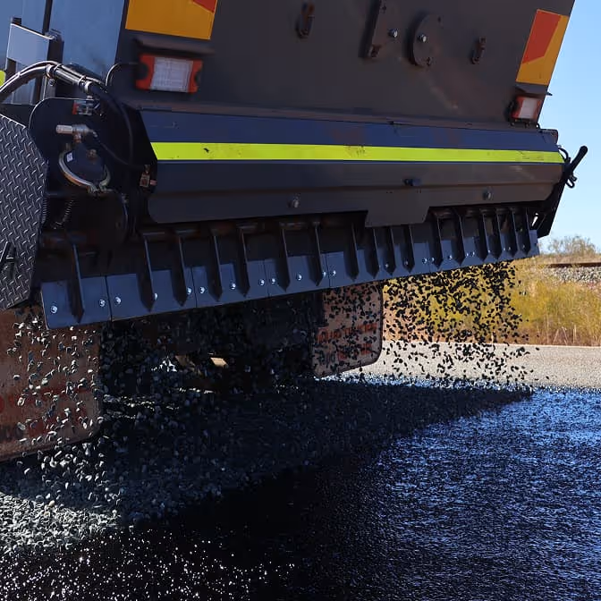 Two dump trucks and a road roller paving asphalt on a remote highway near dry hills and railway tracks.