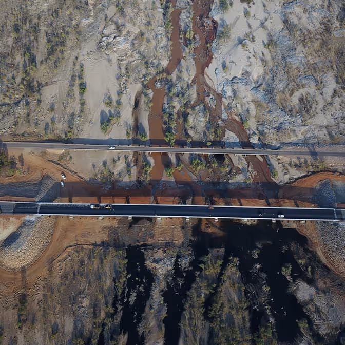Two dump trucks and a road roller paving asphalt on a remote highway near dry hills and railway tracks.