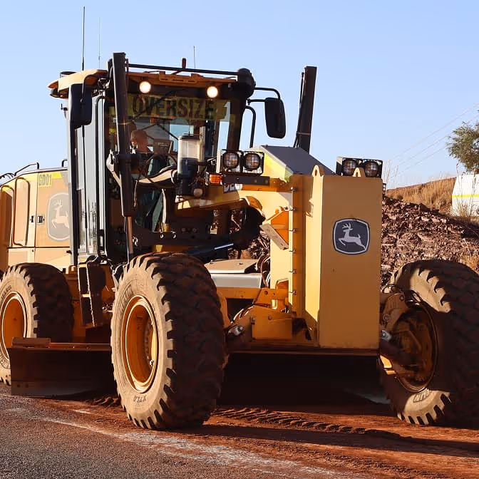 Two dump trucks and a road roller paving asphalt on a remote highway near dry hills and railway tracks.