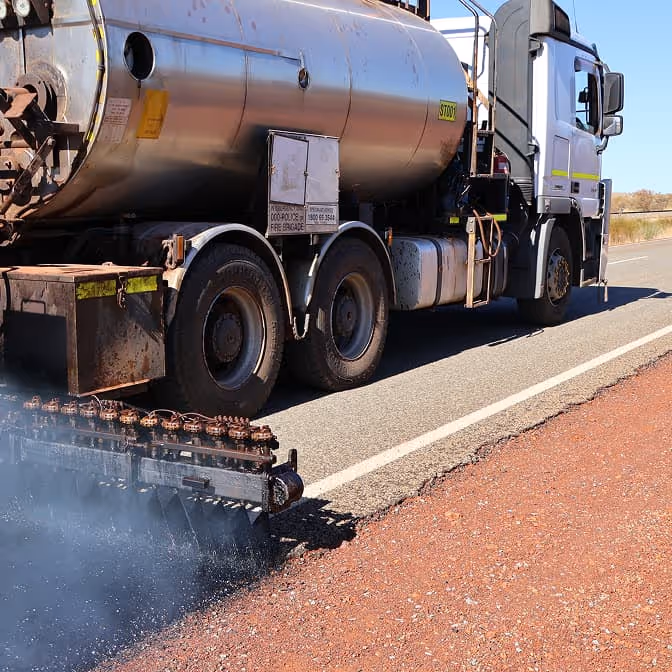 Two dump trucks and a road roller paving asphalt on a remote highway near dry hills and railway tracks.