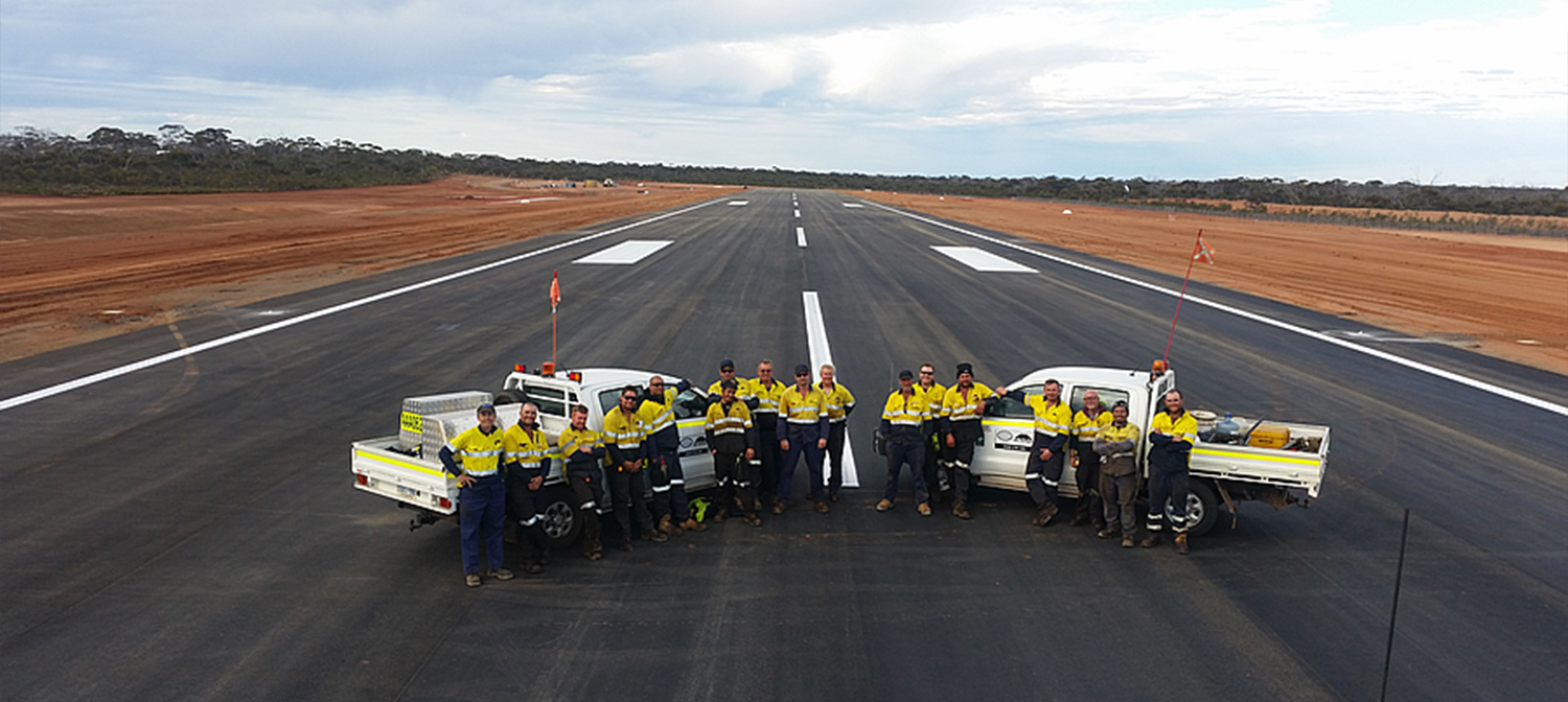 Group of workers in yellow shirts standing between two white utility trucks on a newly paved runway.