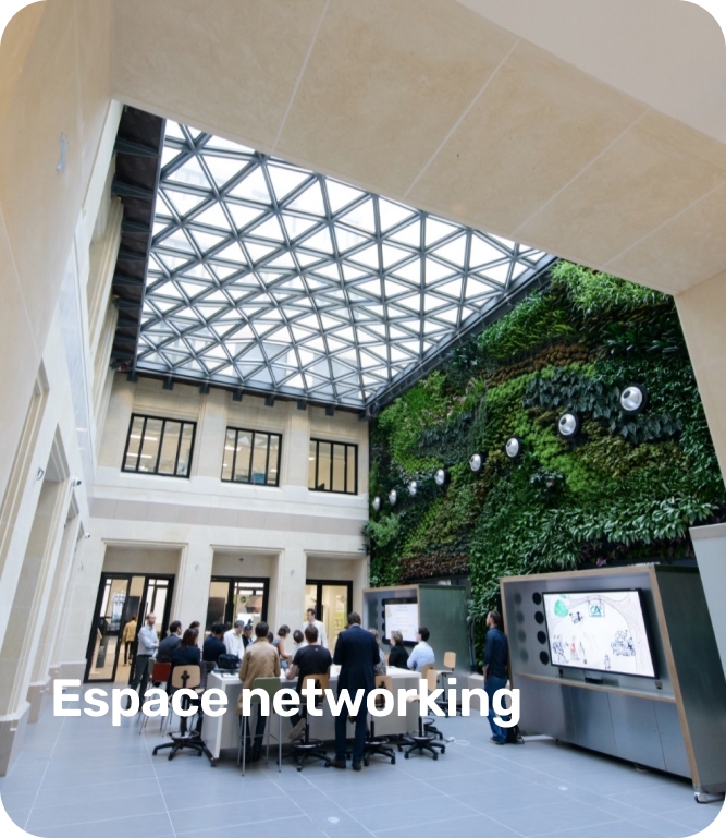 People gathered around tables in a bright indoor networking space with a glass ceiling and a green plant wall.