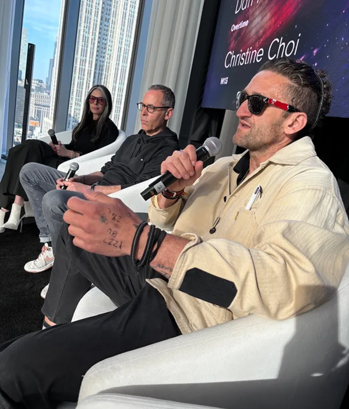 Three people seated in white chairs holding microphones, with city buildings visible through windows behind them.