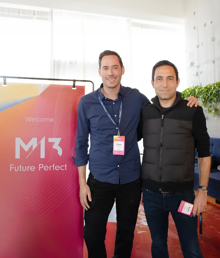 Two men standing side by side smiling, one in a blue shirt and the other in a black vest, near a M13 Future Perfect banner.