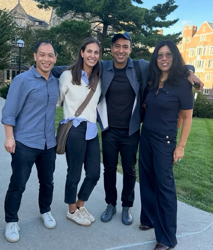 Four adults standing outdoors with arms around each other, smiling on a pathway near grass and buildings.