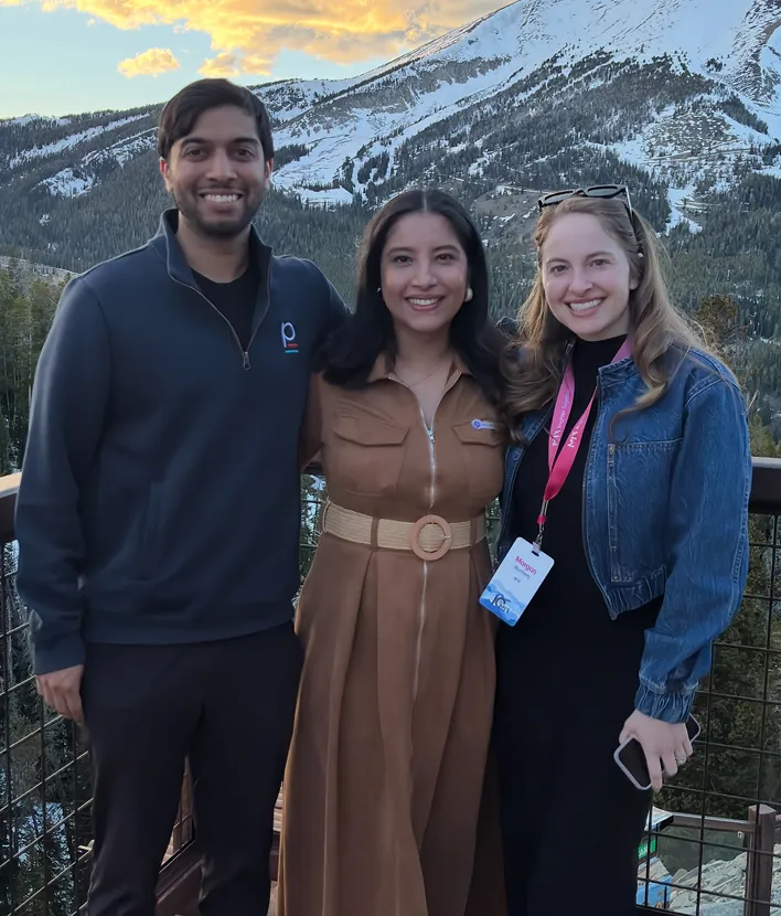 Three people smiling on a mountain balcony with snowy peaks and forest in the background at sunset.