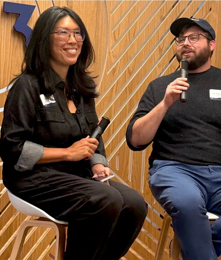 Two people sitting on stools, each holding a microphone and engaged in conversation.
