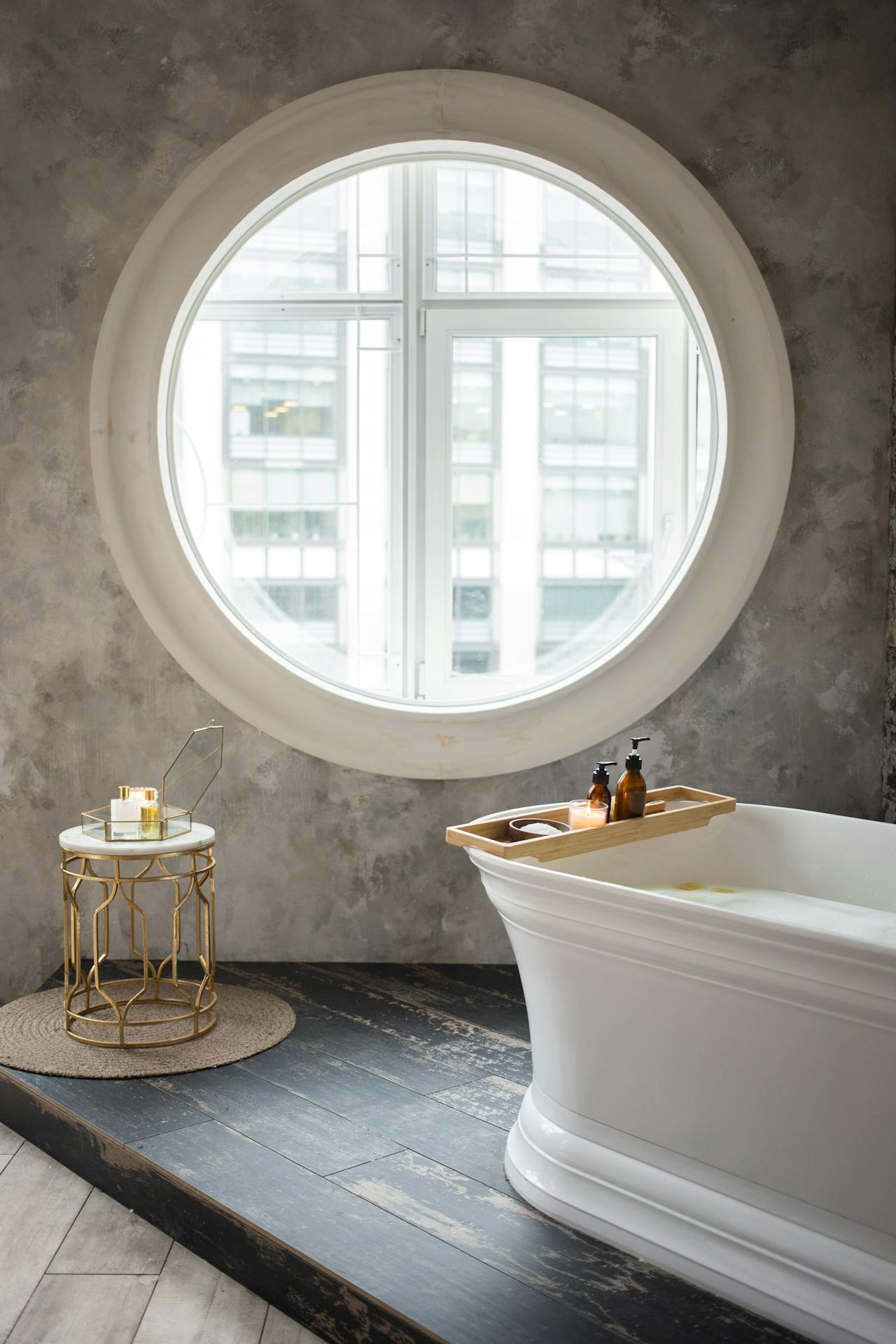 White freestanding bathtub filled with foam beside a round window and a small gold side table with toiletries.