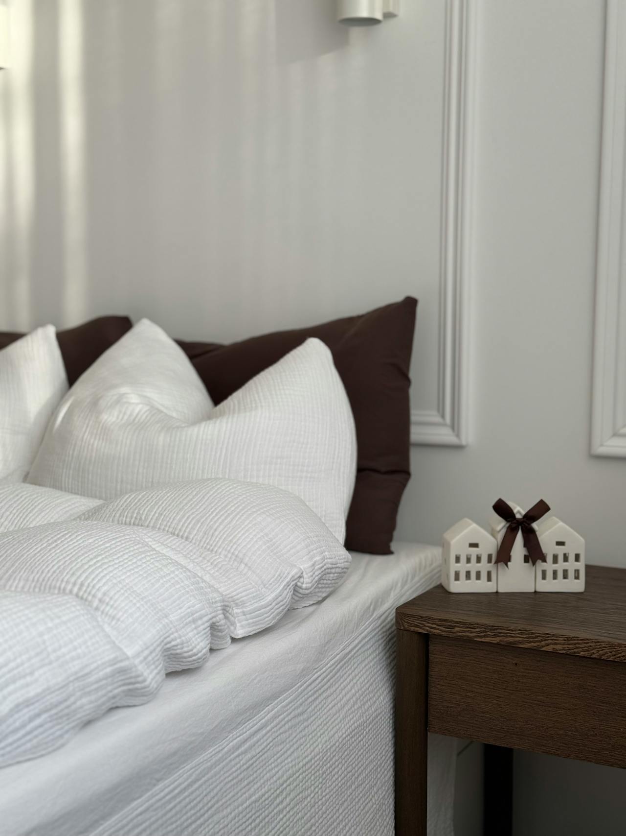 Close-up of a bed with white textured bedding and brown pillows next to a wooden bedside table with small ceramic house decorations tied with a brown ribbon.
