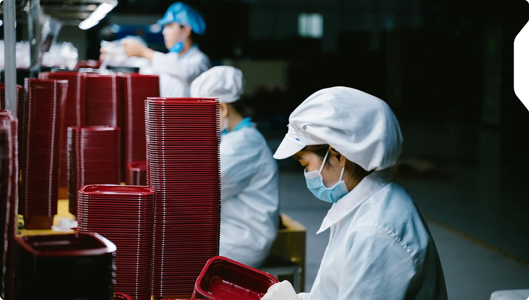 Factory workers in protective clothing and face masks inspecting and stacking red plastic trays.