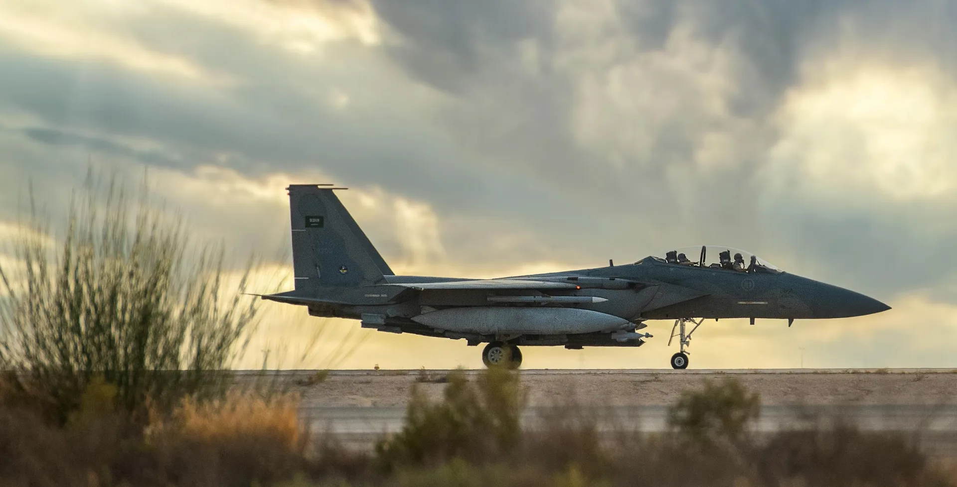 Military fighter jet taxiing on runway at dusk under cloudy sky.