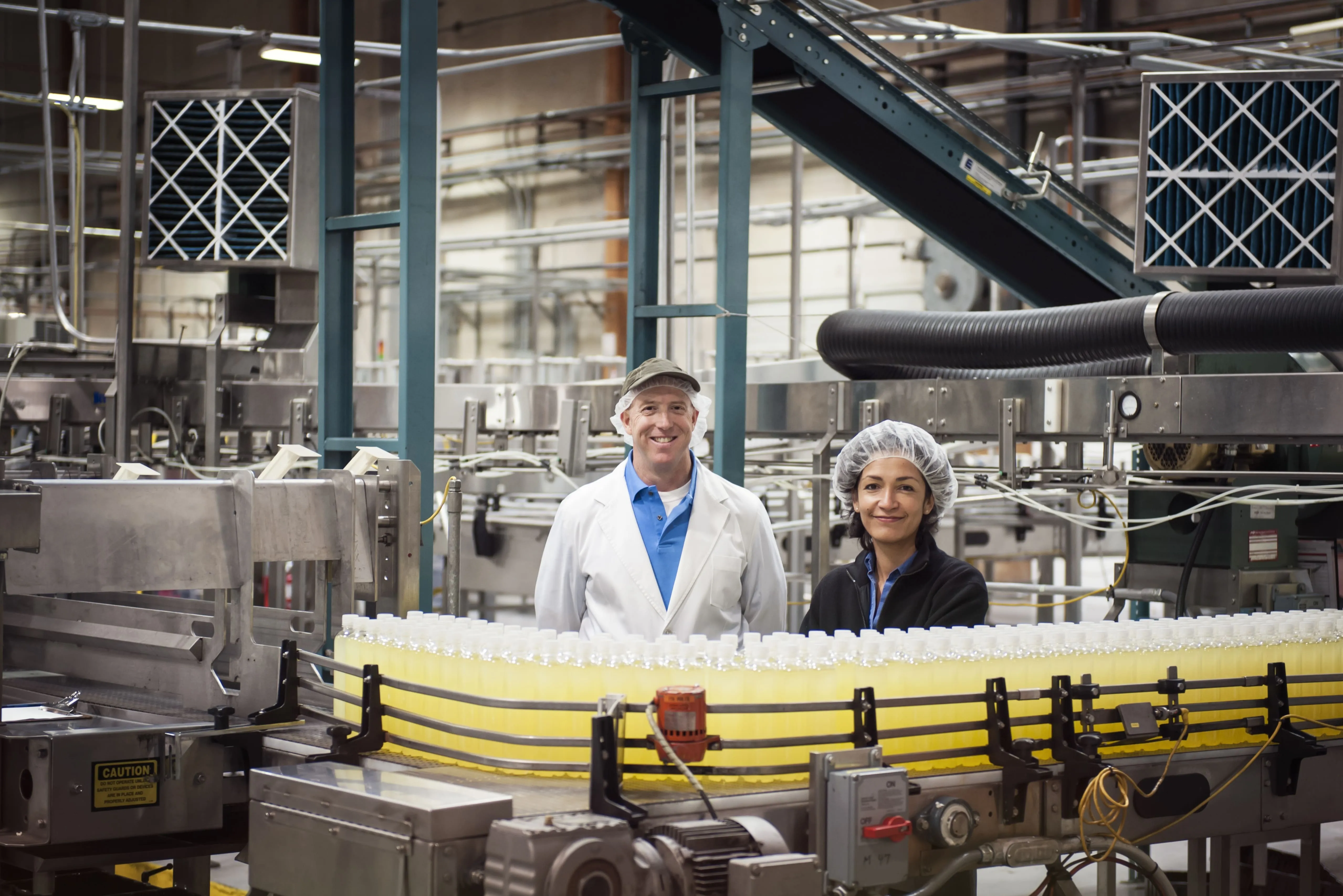 Two factory workers wearing hairnets standing behind a conveyor belt filled with bottles of yellow liquid in a beverage production facility.