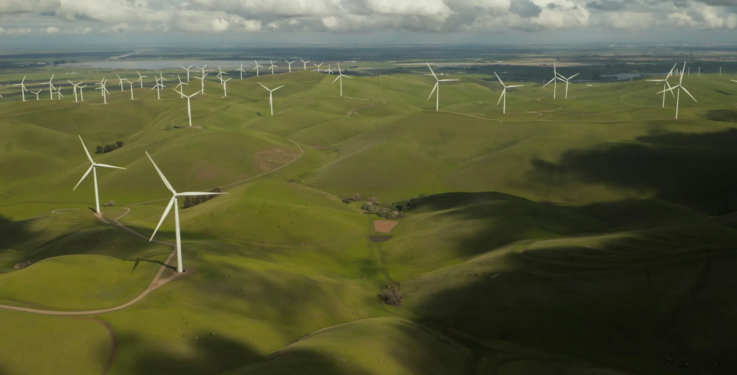 Aerial view of green rolling hills with numerous white wind turbines under a cloudy sky.
