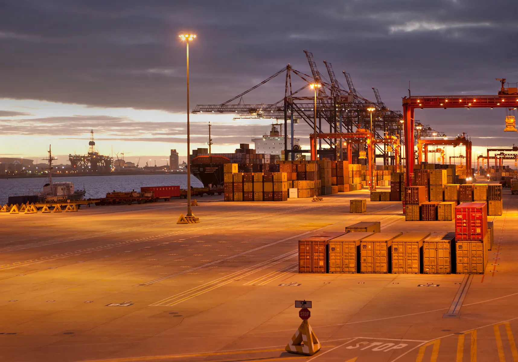 Shipping port at dusk with stacked containers, cranes, and a cargo ship docked near the water.