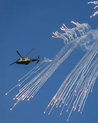 Military helicopter releasing multiple flares creating bright trails against a clear blue sky.