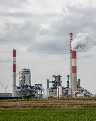 Industrial plant with tall chimneys emitting white smoke under a cloudy sky.