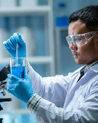 Scientist wearing safety goggles and blue gloves using a pipette to transfer blue liquid in a laboratory beaker.