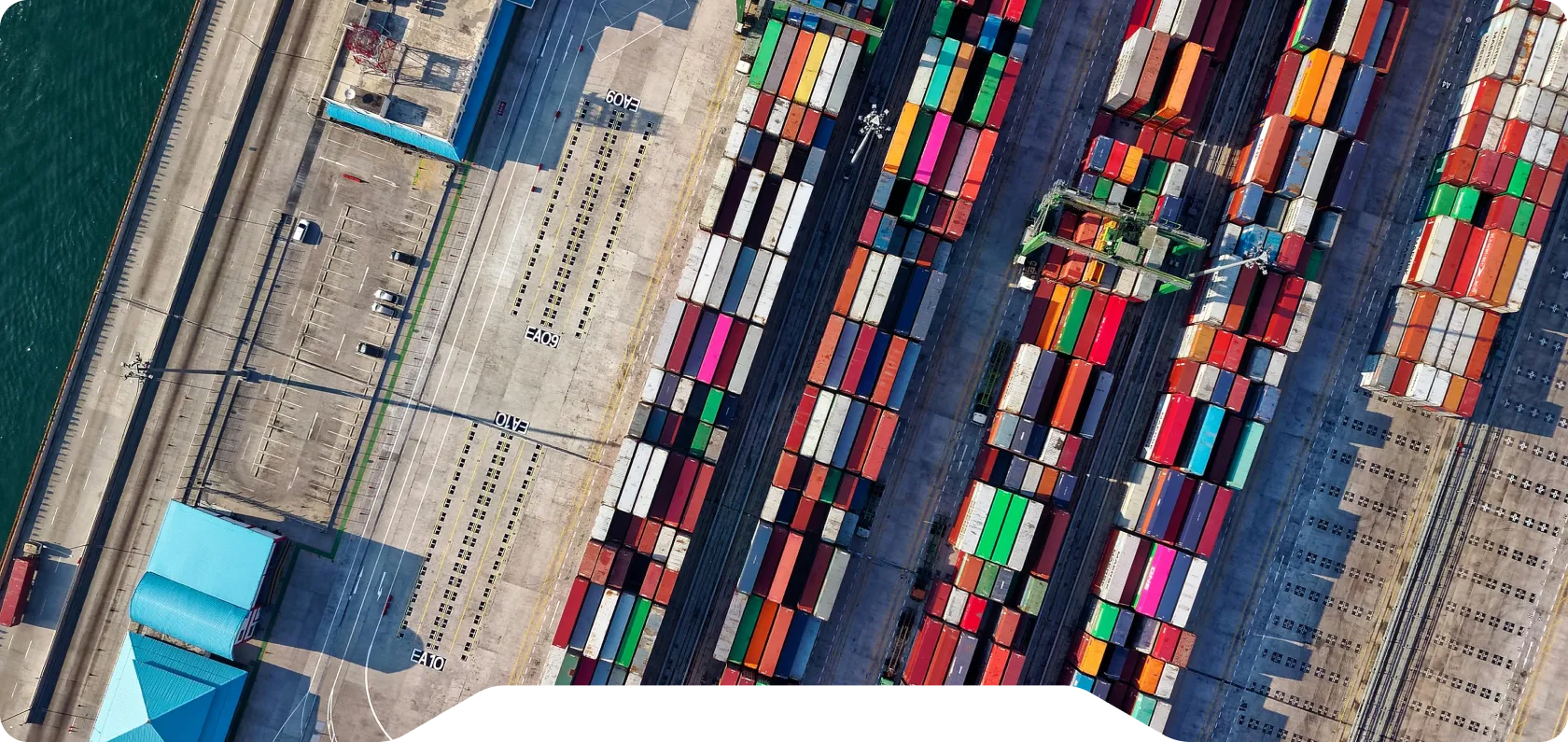 Aerial view of a shipping port with colorful stacked shipping containers and nearby buildings with blue roofs.