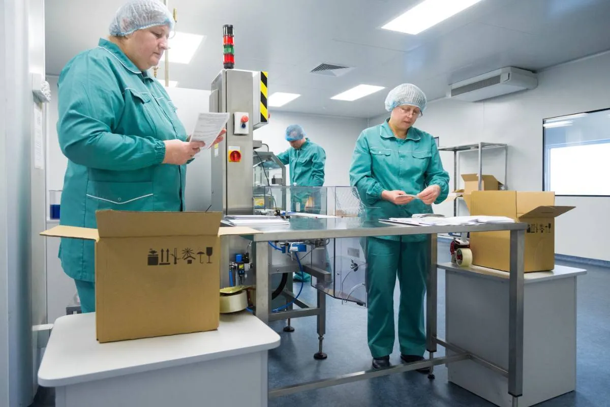 Three factory workers in green uniforms and hairnets inspecting and sorting items at a packaging station with cardboard boxes in a clean production room.