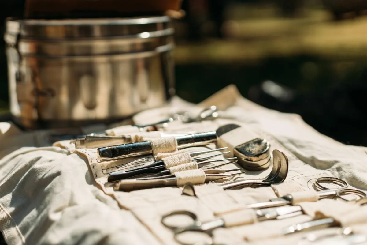 Close-up of vintage surgical or medical tools laid out on a cloth with a metallic container in the background.