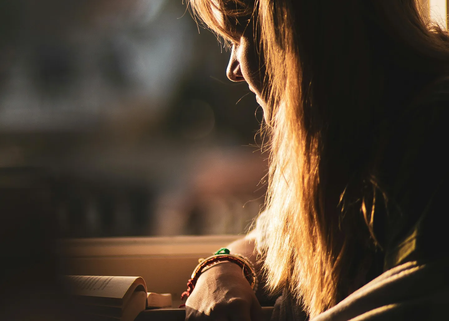 Side view of a woman reading a book in warm sunlight with bracelets on her wrist.