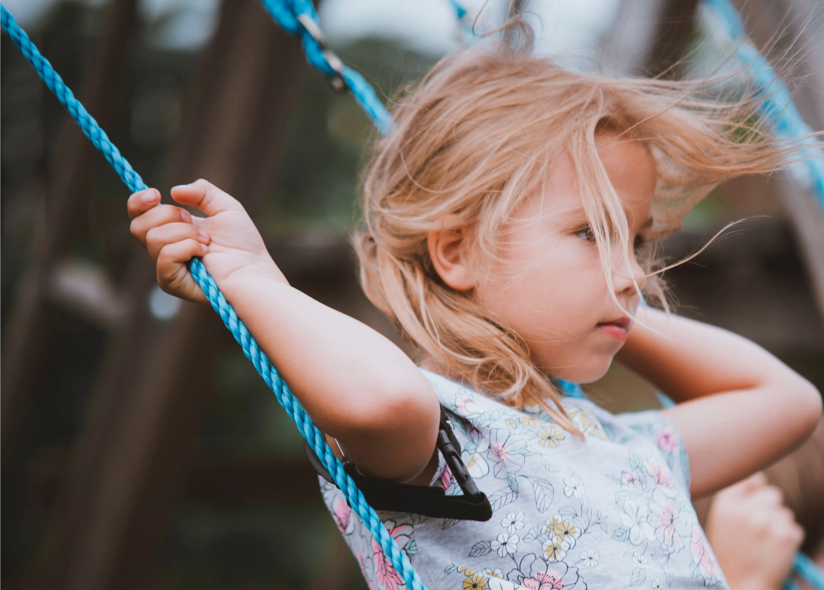 Side view of a blonde child holding blue swing ropes with hair blowing in the wind outdoors.